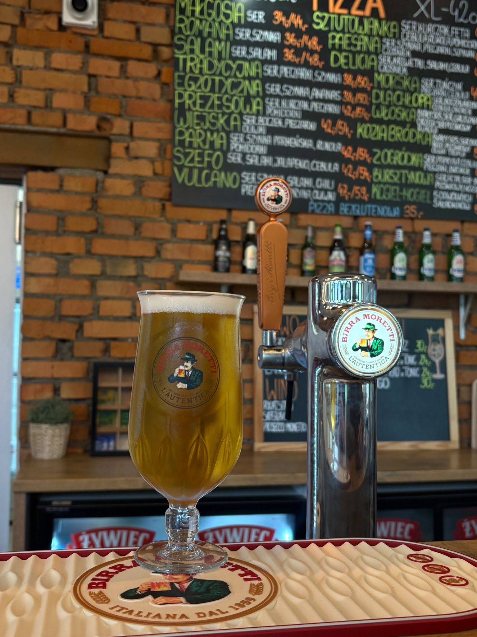 Złociste piwo Birra Moretti z nalewaka w firmowym pokalu, w tle tablica z menu pizzy